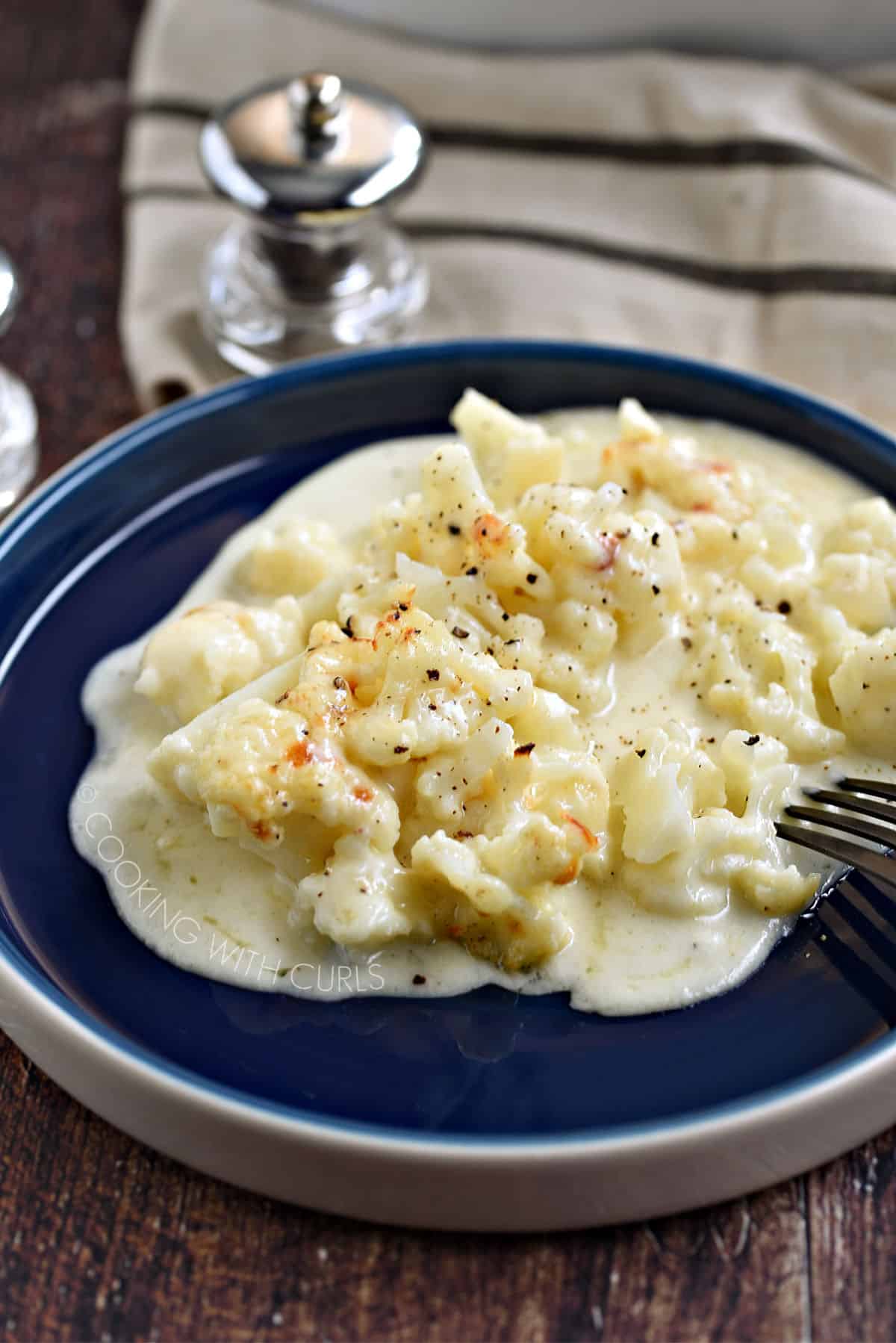 Cauliflower pieces topped with a creamy cheese sauce on a blue plate with a black fork poking in on the right side.