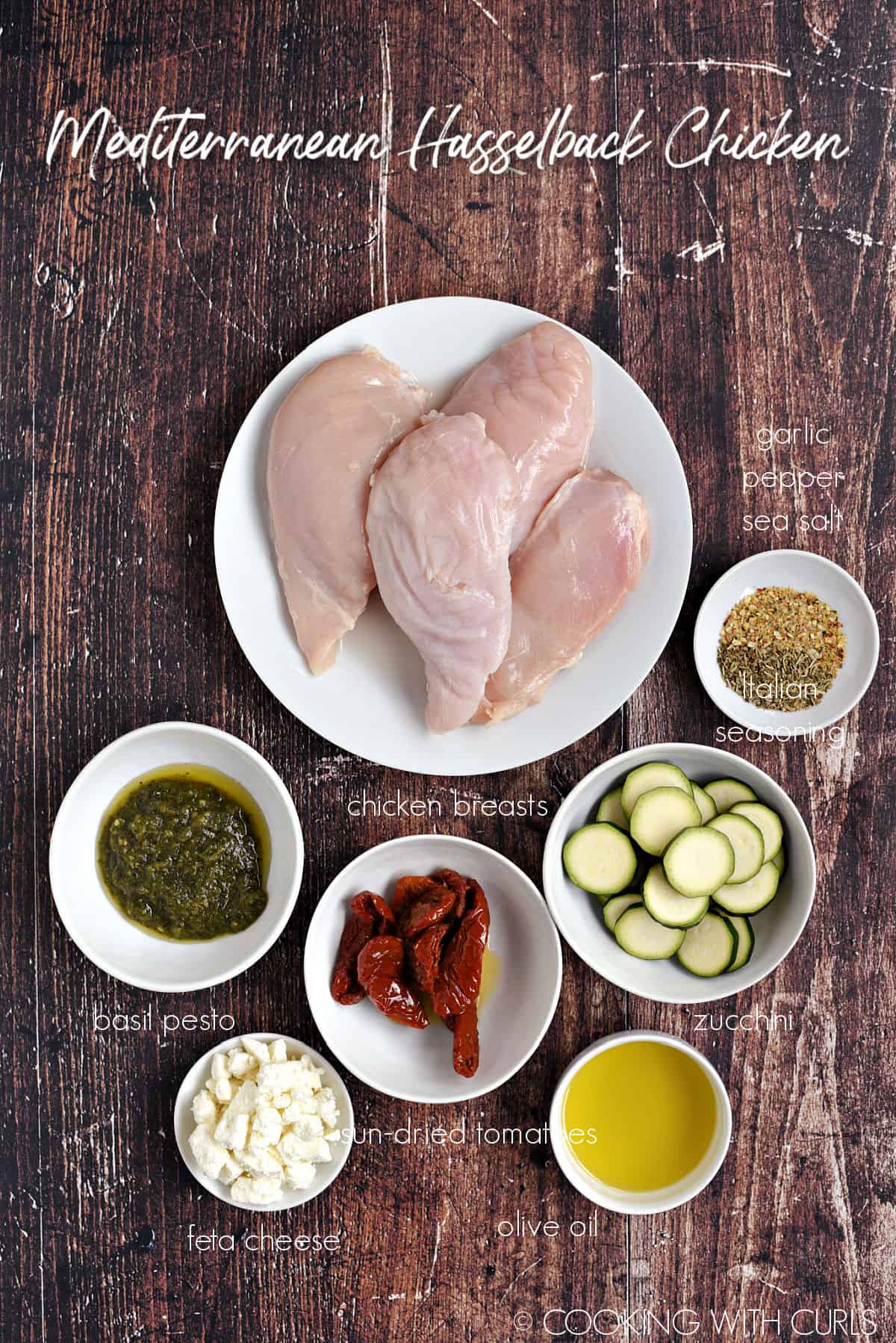 Chicken breasts, basil pesto, sun-dried tomatoes, zucchini rounds, feta cheese, olive oil, Italian seasoning and garlic-pepper sea salt in white bowls.
