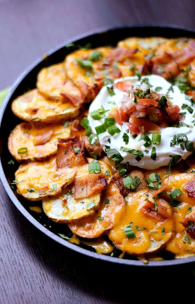 Sliced potato nachos topped with melted cheese, bacon, sour cream, salsa, and green onion in a cast iron skillet.