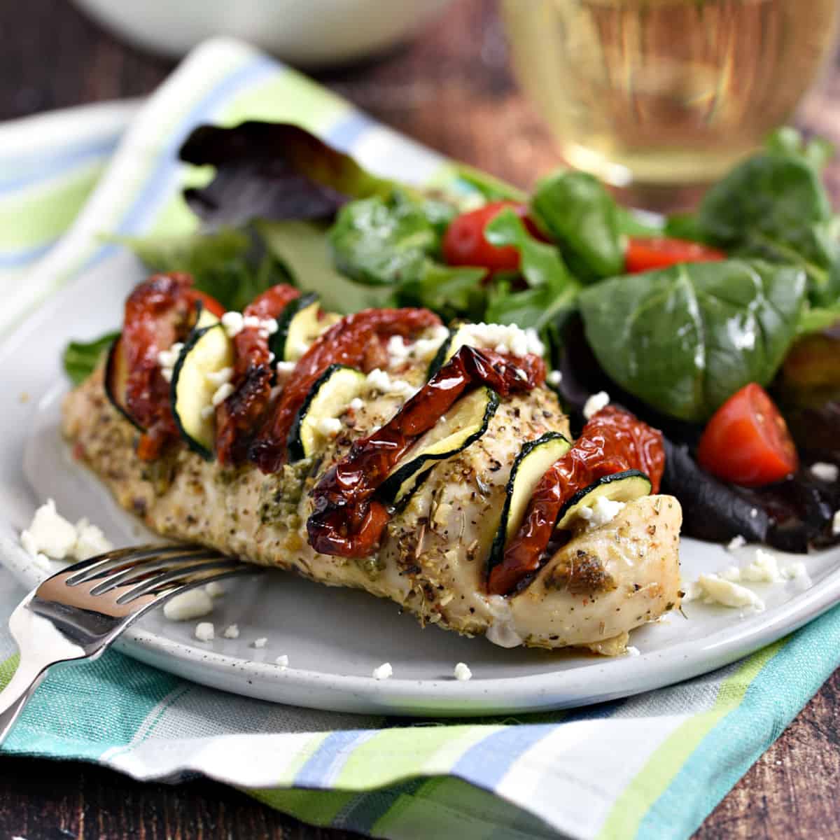 Chicken breast stuffed with pesto, sun-dried tomatoes and zucchini slices on a plate with mixed greens and cherry tomatoes.