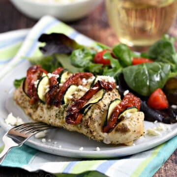 Chicken breast stuffed with pesto, sun-dried tomatoes and zucchini slices on a plate with mixed greens and cherry tomatoes.