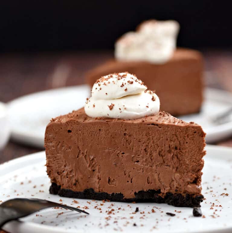A slice of chocolate cheesecake topped with whipped cream and sprinkled with shaved chocolate sitting on a white plate with a second slice in the background.