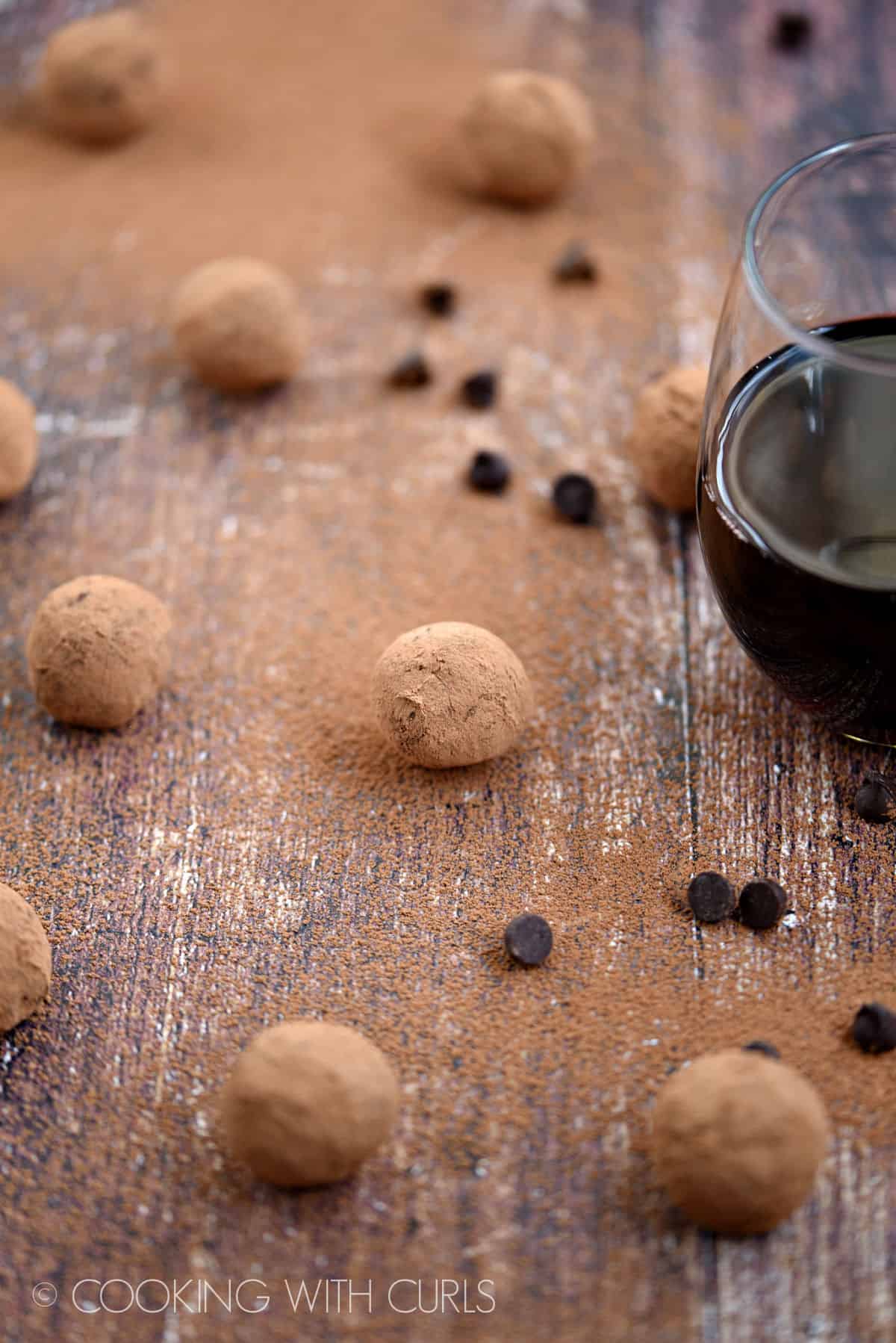 Cocoa covered red wine chocolate truffles on a wooden surface surrounded by cocoa powder, chocolate chips and a glass of wine.