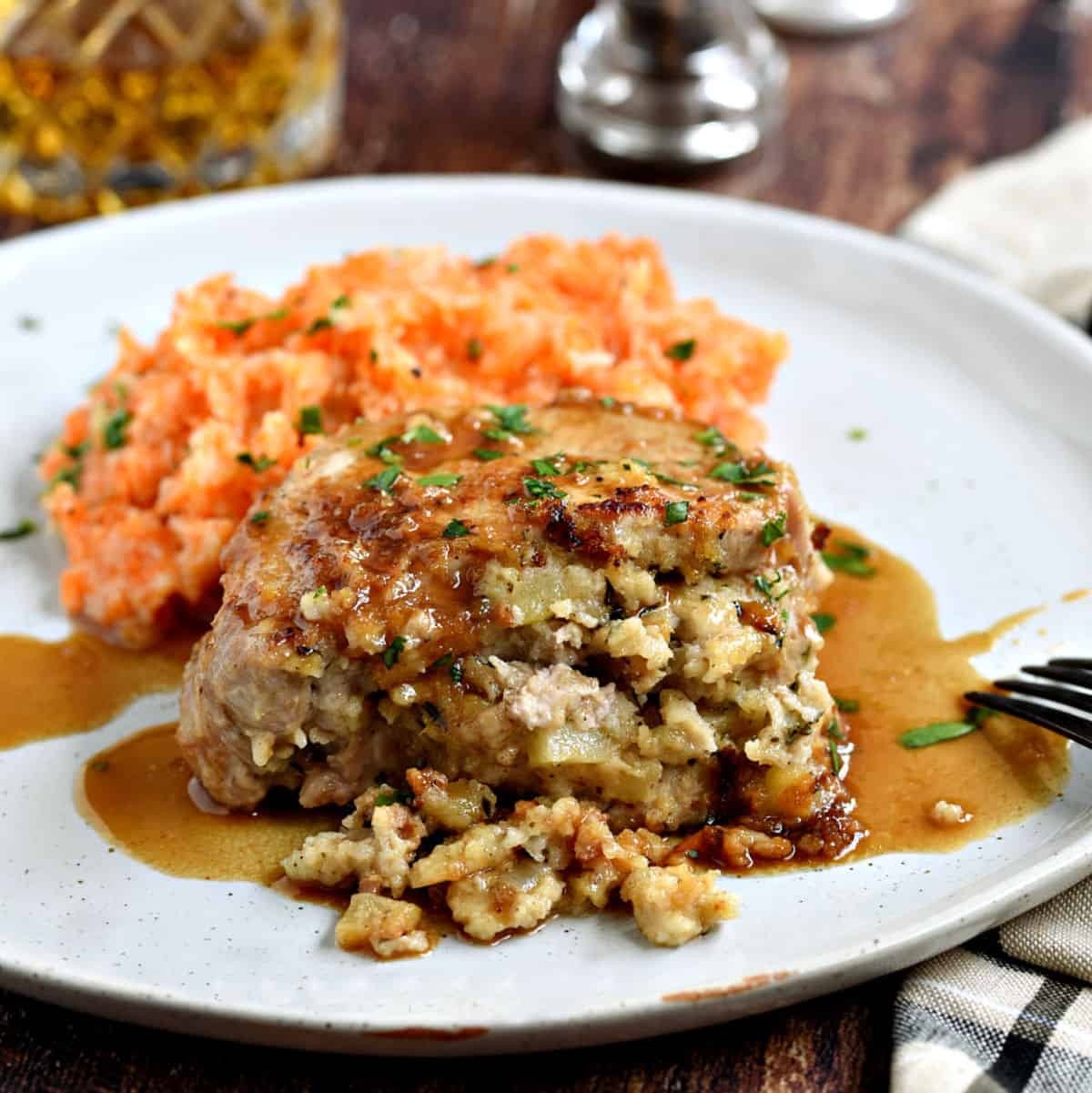 A stuffed pork chop covered in an apple-bourbon sauce and served with carrot parsnip mash on a large white plate with a glass of whisky in the background.