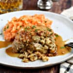 A stuffed pork chop covered in an apple-bourbon sauce and served with carrot parsnip mash on a large white plate with a glass of whisky in the background.