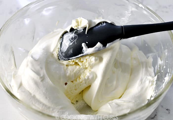 Whipped cream being folded into the cream cheese mixture in a glass bowl with a gray silicone spatula. 