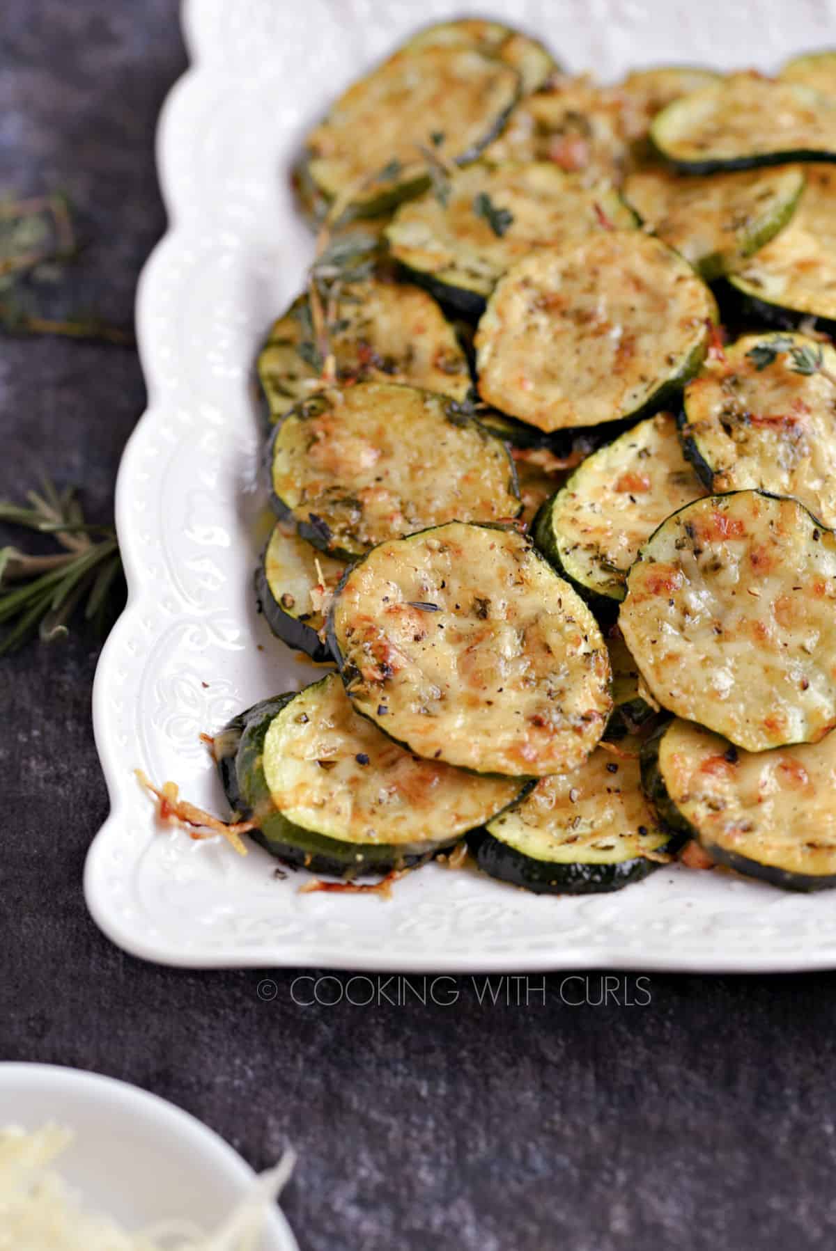 Melted cheese topped zucchini slices stacked on a white platter with sprigs of fresh rosemary scattered around and a small bowl of grated Parmesan.