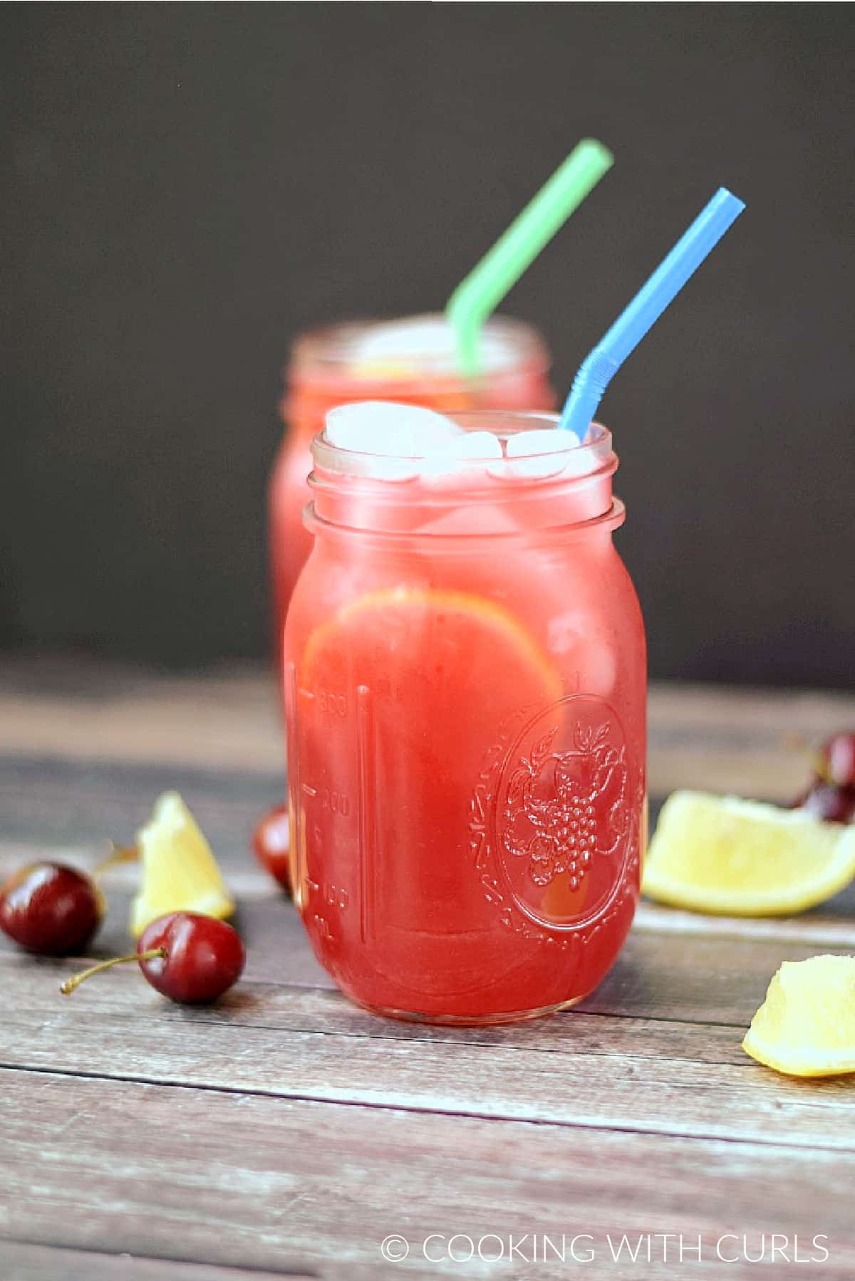 Two glass mason jars filled with cherry lemonade, ice and lemon slices with fresh cherries and lemon wedges scattered on the board.