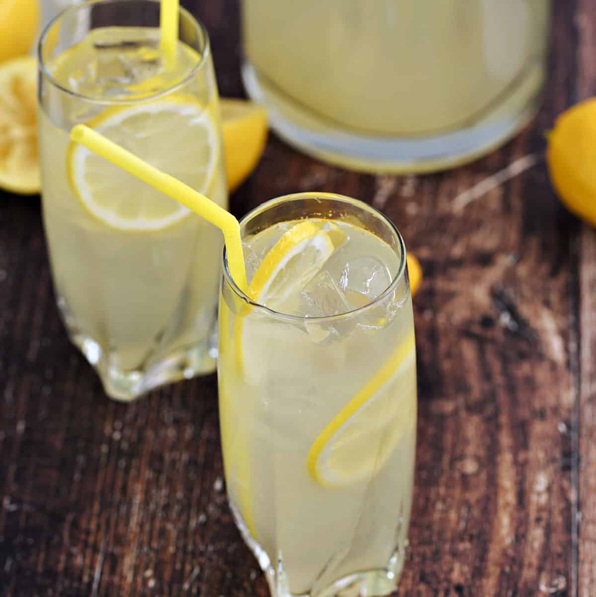 Two glasses and one glass pitcher filled with lemonade and lemon slices and yellow straws.