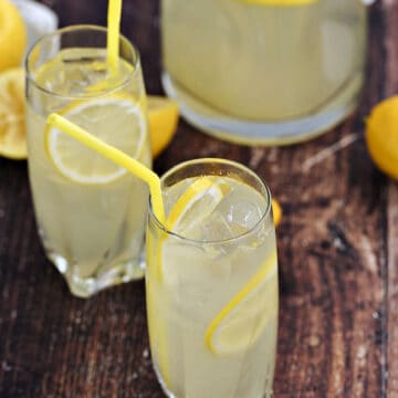 Two glasses and one glass pitcher filled with lemonade and lemon slices and yellow straws.