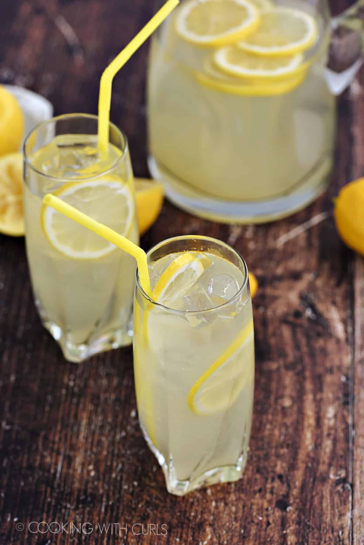 Two, tall glasses and a glass pitcher filled with ice, lemonade, and lemon slices with yellow straws.