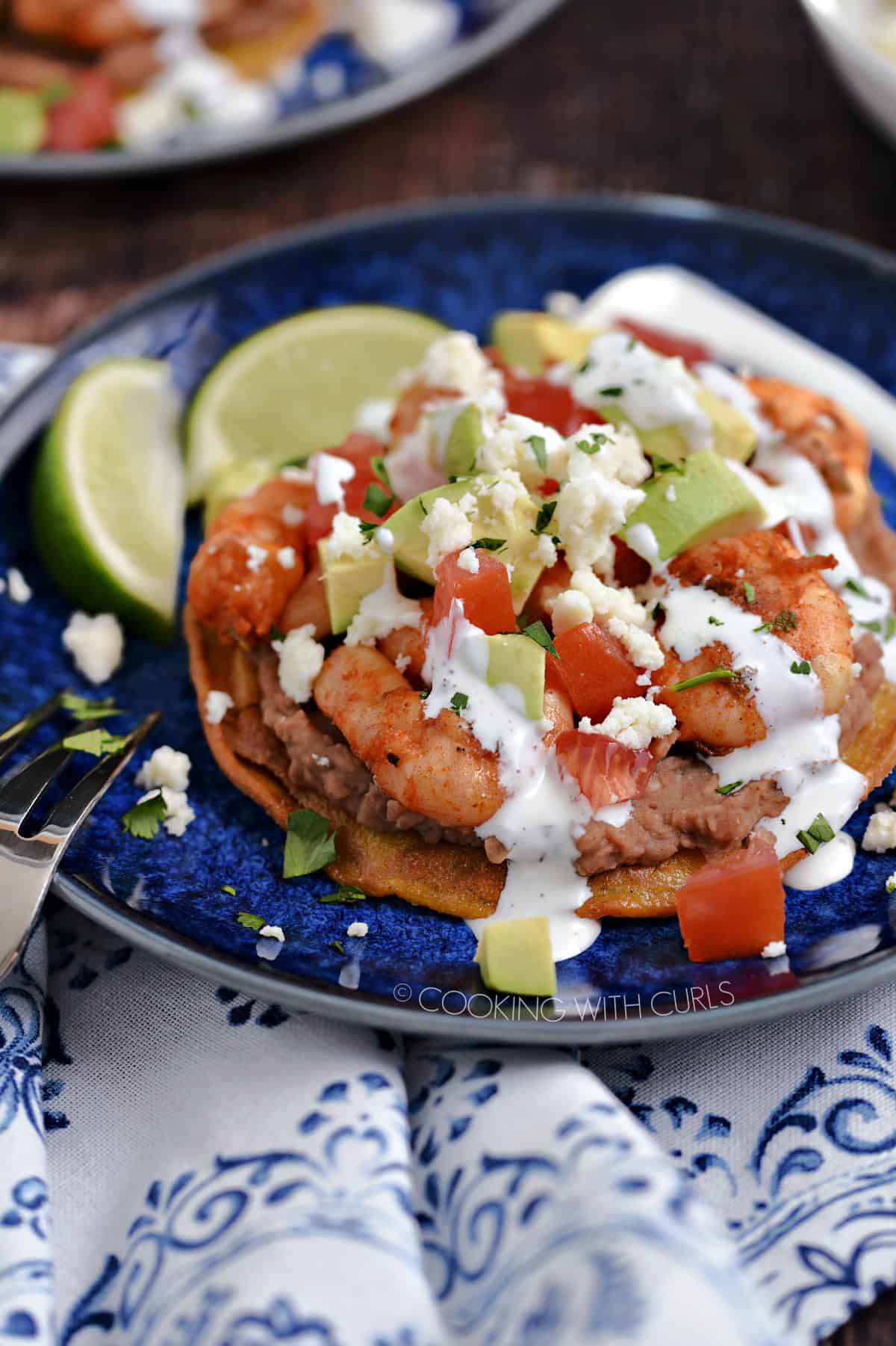 Shrimp, diced tomatoes, onions and avocado on a bed of refried beans on a corn tortilla drizzled with crema and crumbled cheese on a blue plate.
