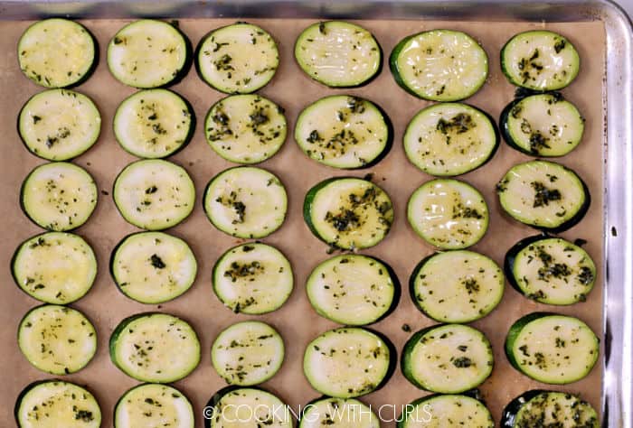 Herb butter brushed on top of zucchini slices on a parchment lined baking sheet. 