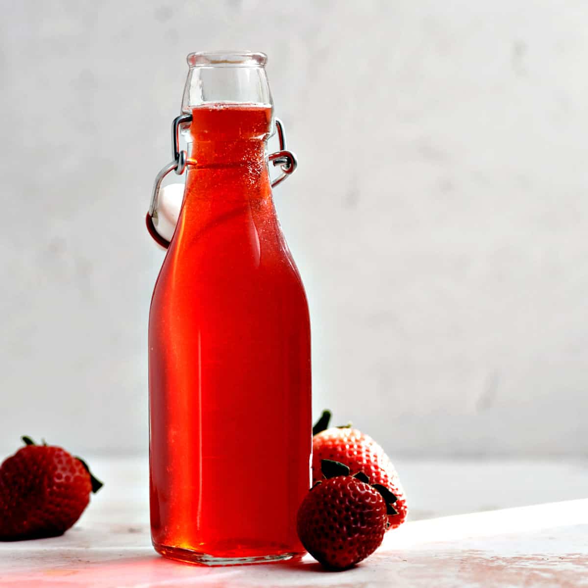Strawberry Simple Syrup in a square bottle with three fresh strawberries around the base of the bottle.