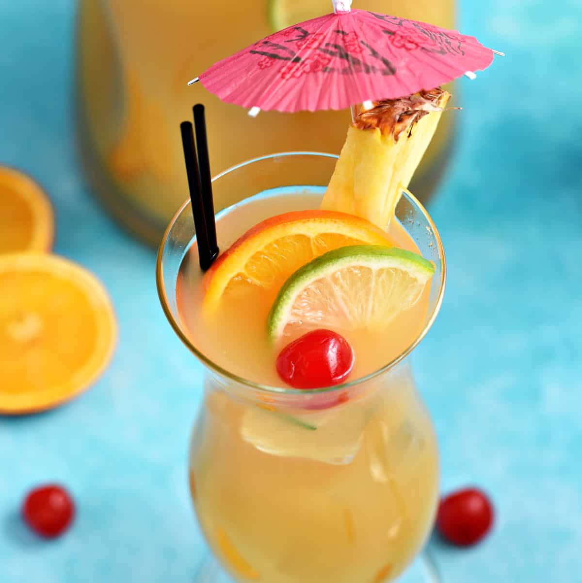 A hurricane glass filled with Jamaican Rum Punch and garnished with slices of orange and lime with a cherry and a pineapple slice with a pink paper umbrella on the rim.