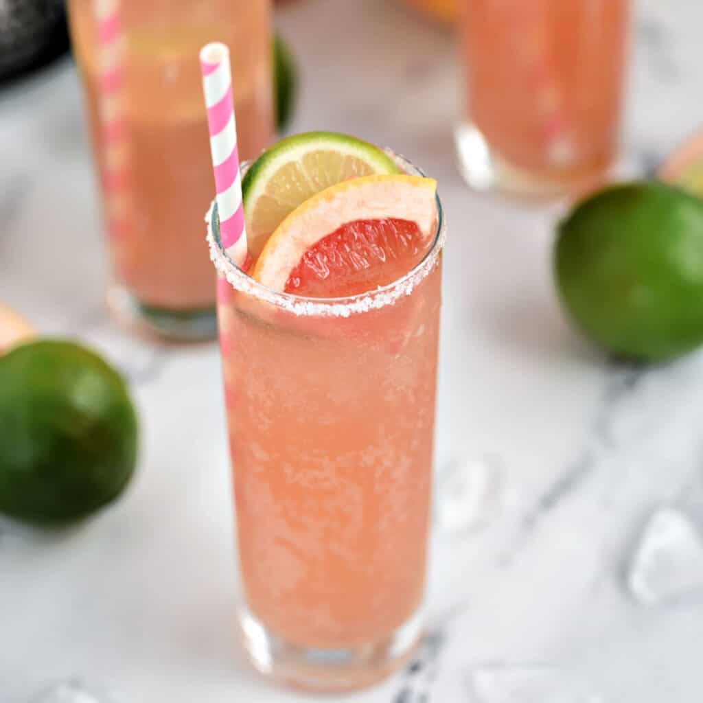 A bubbly, pink cocktail in a tall glass garnished with a slice of lime and grapefruit and a pink and white striped straw.