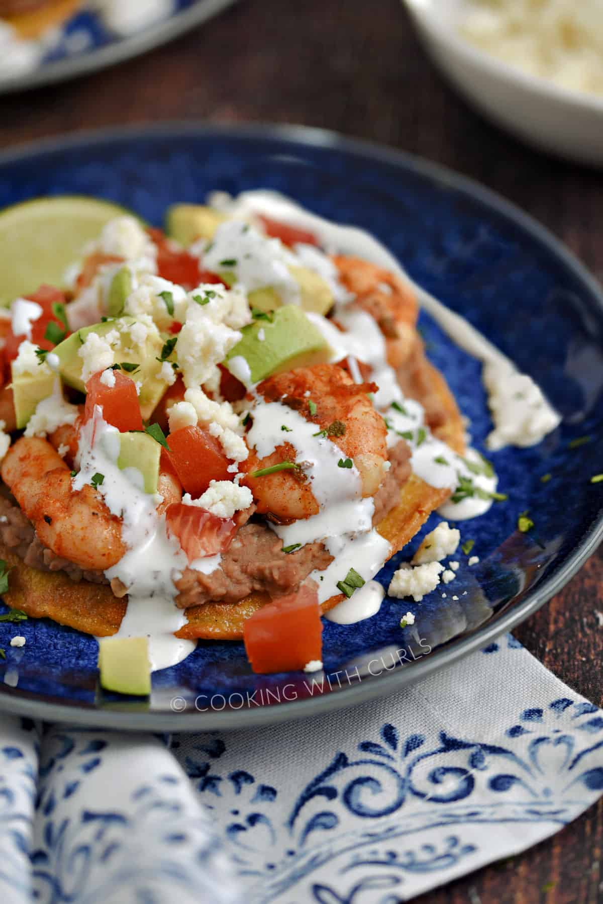 Close-up image of seasoned shrimp, diced tomatoes, onions and avocado on a bed of refried beans on a corn tortilla drizzled with crema and crumbled cheese on a blue plate.