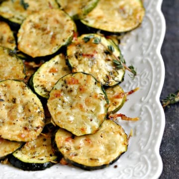 Melted cheese topped zucchini slices stacked on a white platter.