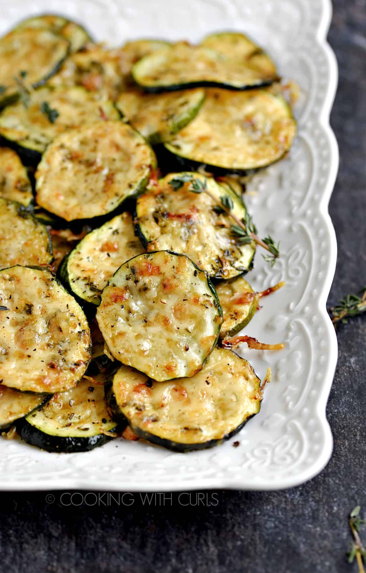 Melted cheese topped zucchini slices stacked on a white platter with sprigs of fresh thyme scattered around.