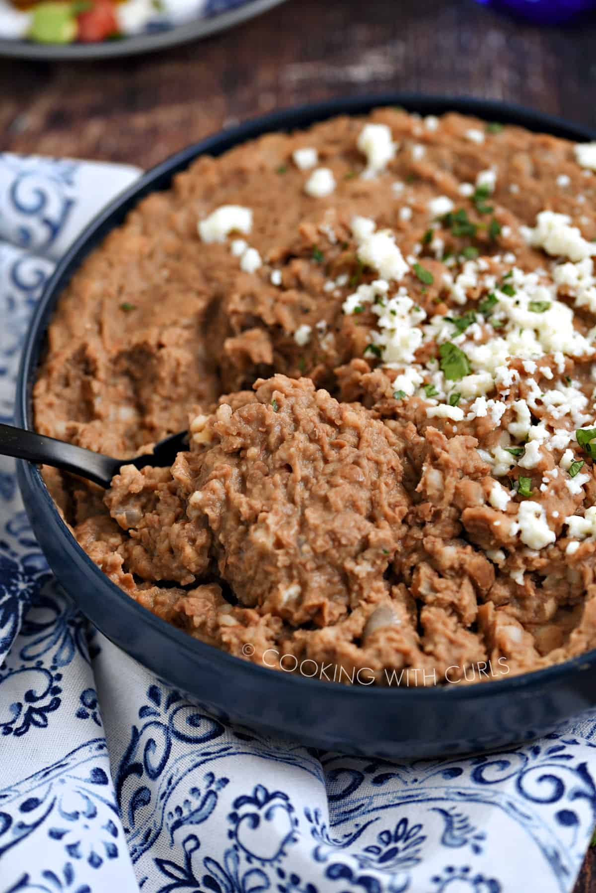 Creamy, mashed Instant Pot refried beans in a blue bowl topped with crumbled cheese and chopped cilantro being scooped with a large black spoon.