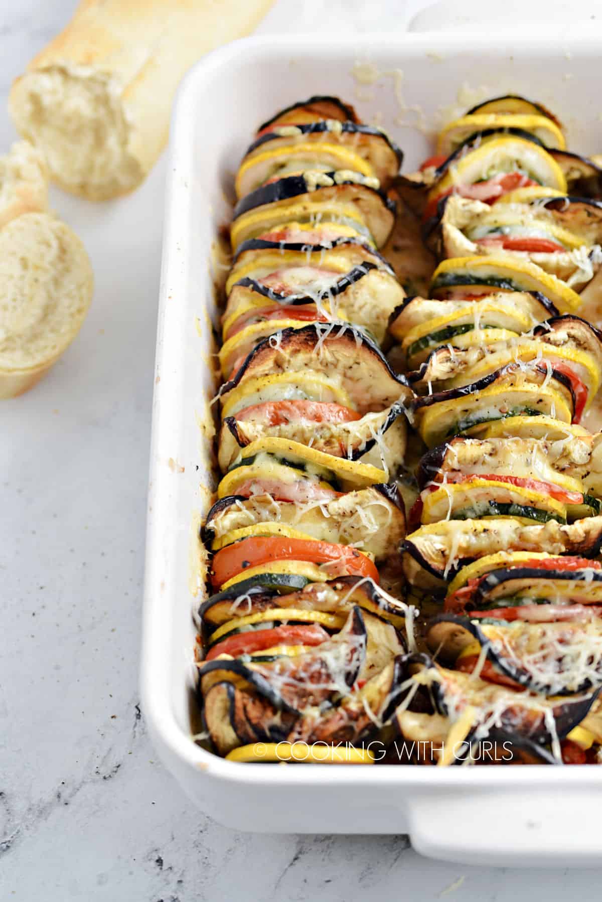 Slices of zucchini, yellow squash, eggplant and tomatoes lined up in a white baking dish topped with melted cheese with French bread in the background.