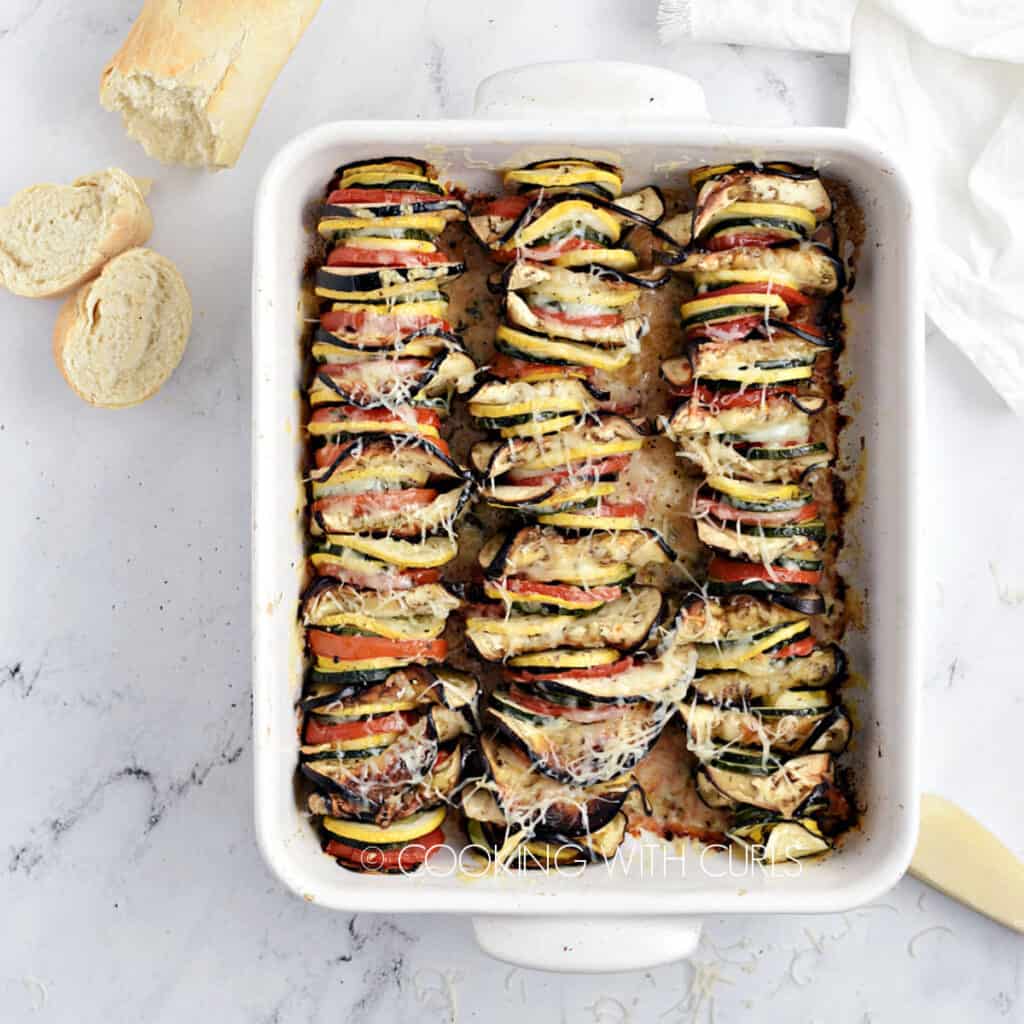 Slices of zucchini, yellow squash, eggplant and tomatoes lined up in a white baking dish topped with melted cheese.