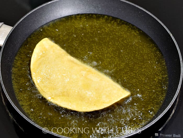 Corn tortilla folded in half in boiling oil inside a small skillet. 