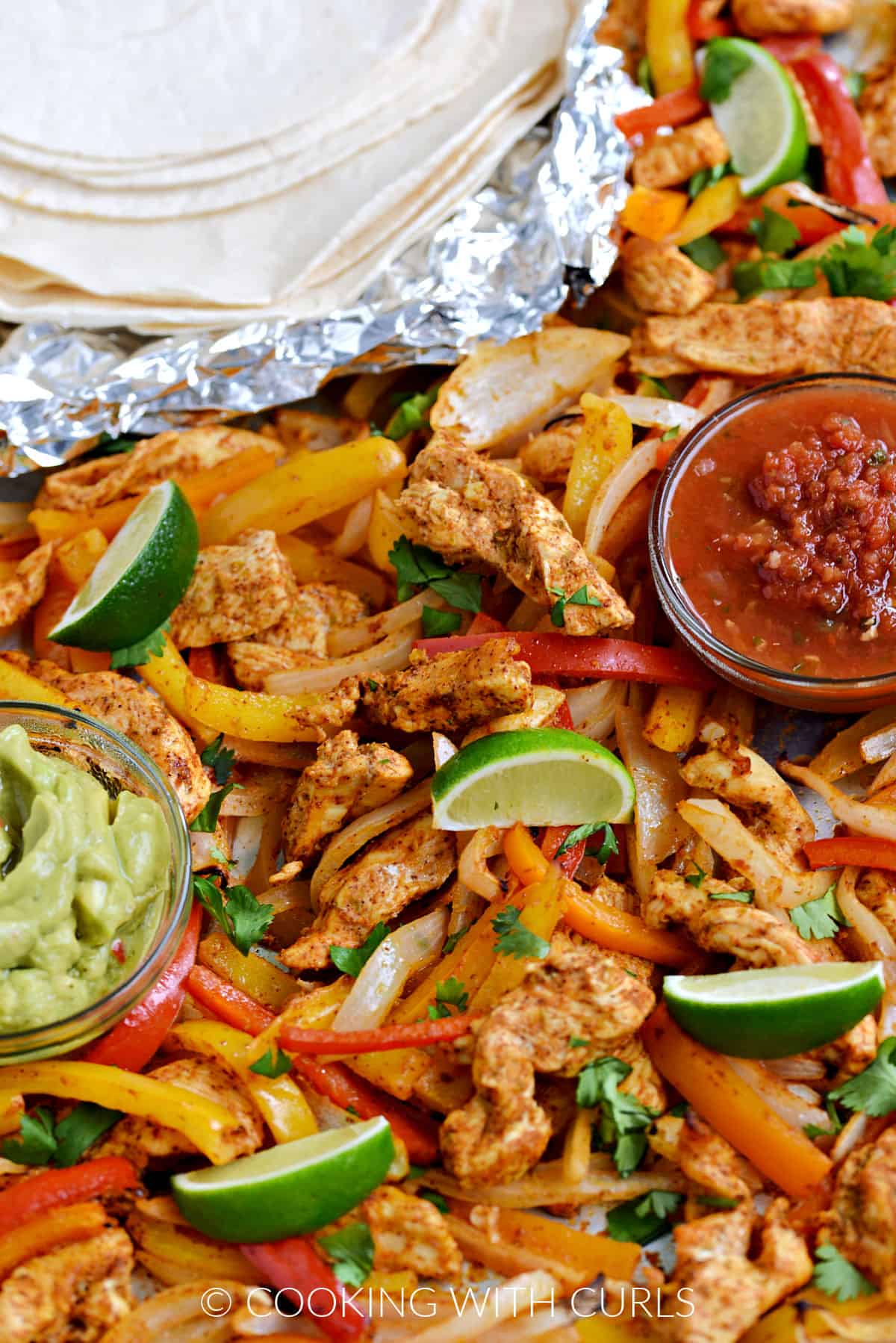 Strips of chicken, onion, red, orange and yellow bell peppers on a sheet pan with bowls of guacamole and salsa, and flour tortillas in the upper left corner.