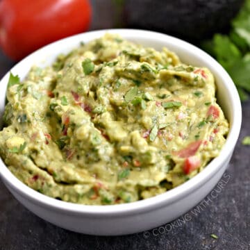 a bowl of guacamole with a lime, tomato, garlic cloves and cilantro leaves in the background.