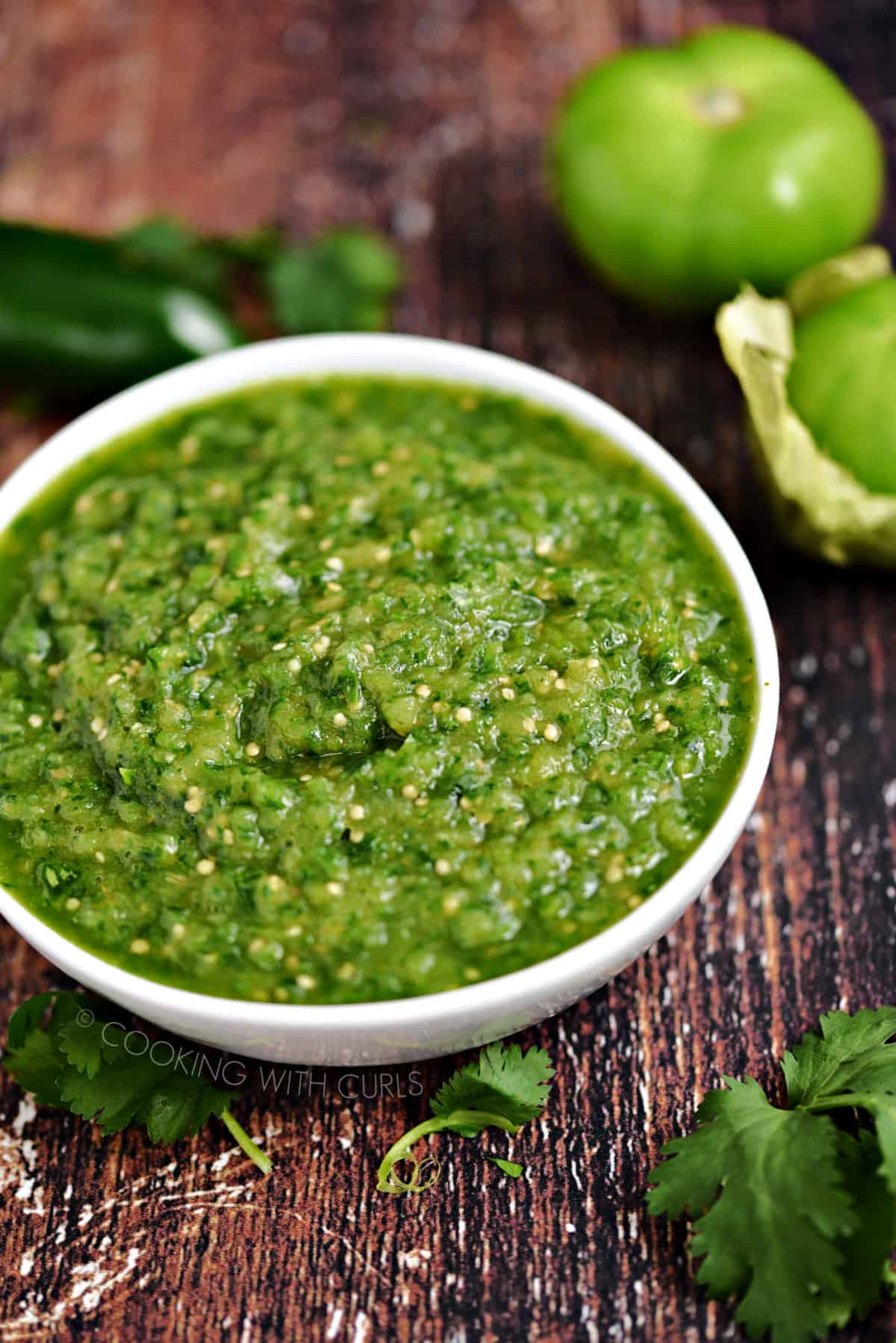 Blended, Instant Pot Salsa Verde in a white bowl surrounded by cilantro, tomatillos and a jalapeno pepper.