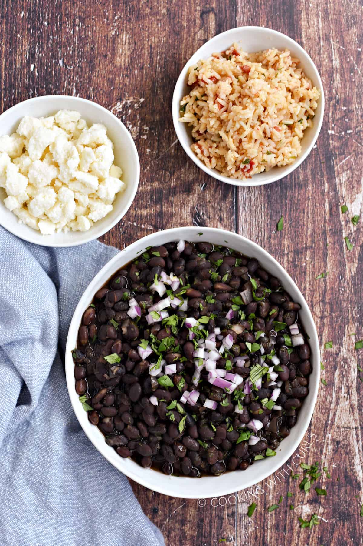 Looking down on black beans in a white bowl topped with diced red onion and chopped cilantro, with cowls of crumbled queso and rice in the background.