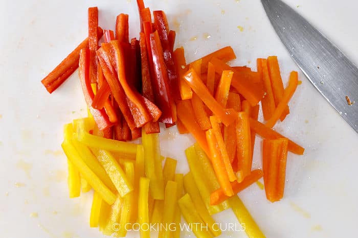 Red, orange and yellow bell peppers cut into strips with a chefs knife on a white cutting board. 
