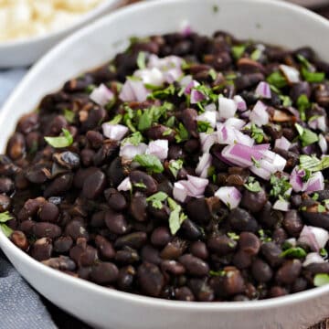 Black beans in a white bowl topped with diced red onion and chopped cilantro with small bowls of rice and crumbled queso in the background..