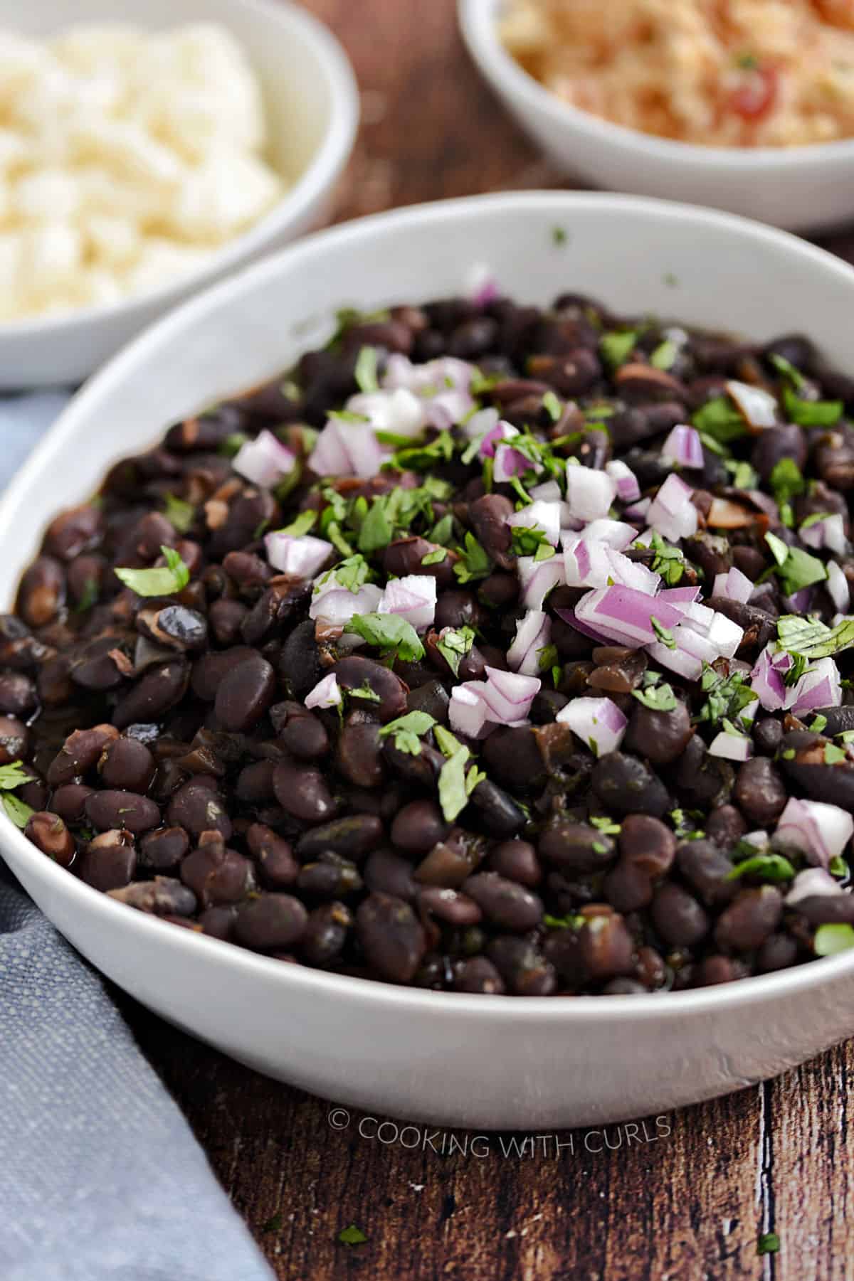 Black beans in a white bowl topped with diced red onion and chopped cilantro, with cowls of crumbled queso and rice in the background.