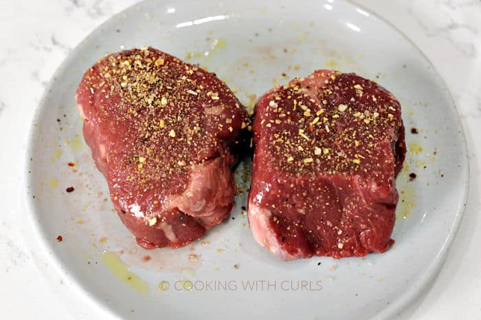 Two steaks coated in oil and steak seasoning on a white plate. 