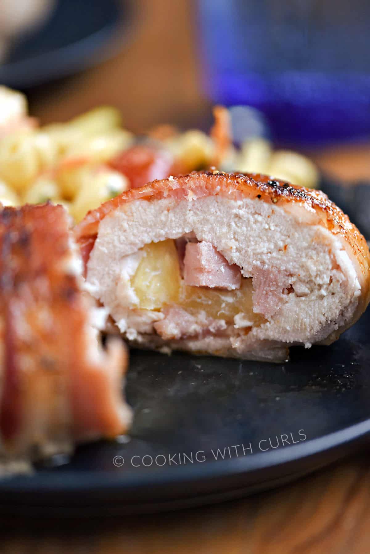 A bacon wrapped chicken breast surrounding chopped ham and pineapple cut in half on a blue plate. 