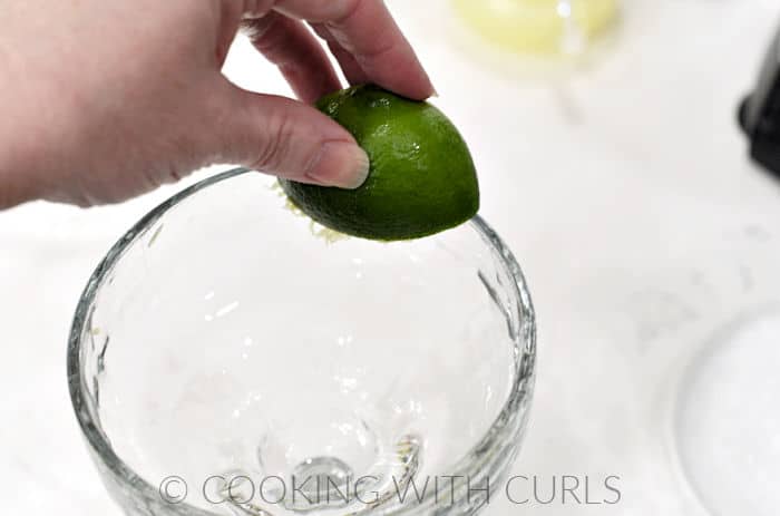 A hand rubbing a squeezed lime around the edge of a margarita glass.