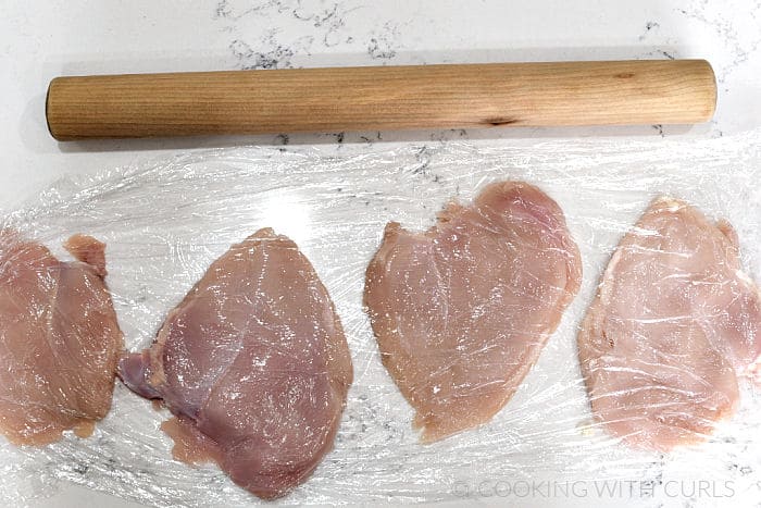 Four chicken breasts flattened between two pieces of plastic wrap by a rolling pin. 