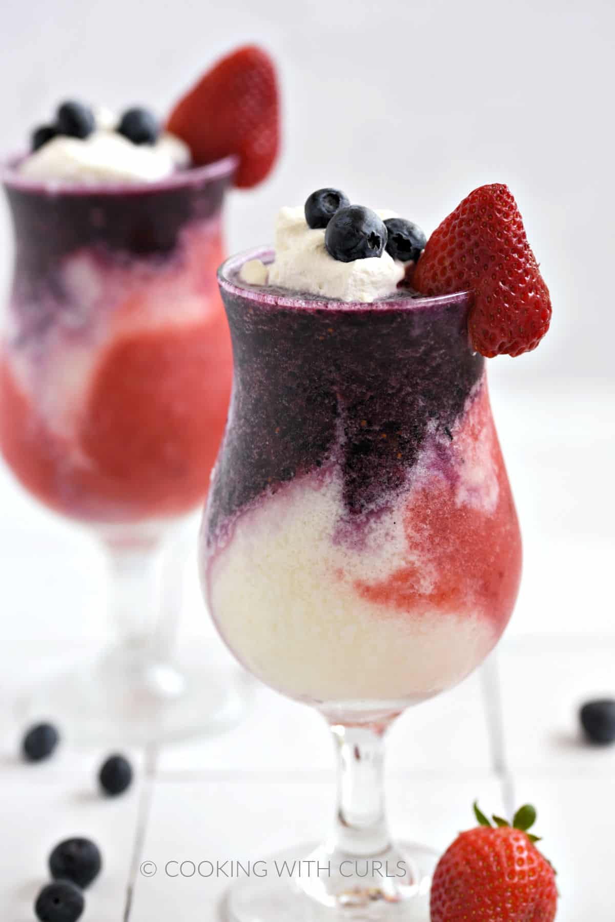 Blueberry, white, and strawberry layers topped with whipped cream, fresh blueberries and a strawberry in two hurricane glasses with scattered berries around the base.