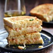 Two halves of a shrimp grilled cheese stacked on top of each other on a blue plate with a second sandwich and glass of white wine in the background.