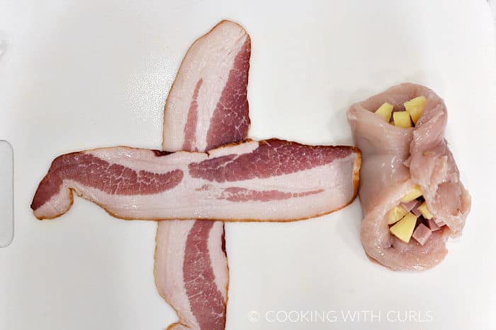 Two slices of bacon laying in a cross pattern on a white cutting board next to a chicken breast wrapped around pineapple and ham chunks. 