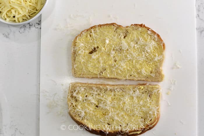 Two slices of bread topped with softened butter and grated Parmesan cheese laying on a white cutting board. 
