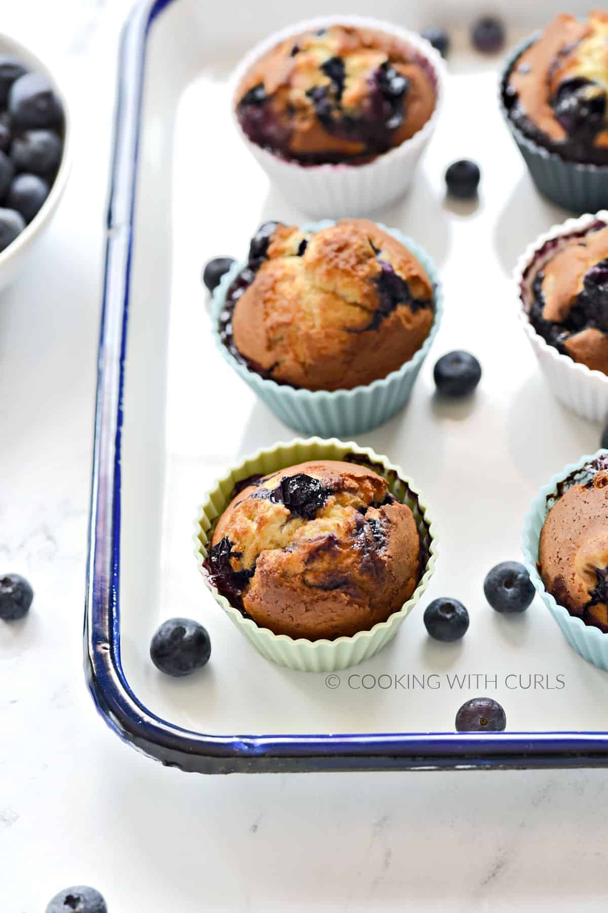 Six blueberry muffins in silicone muffin cups sitting on a white tray surrounded by fresh blueberries.