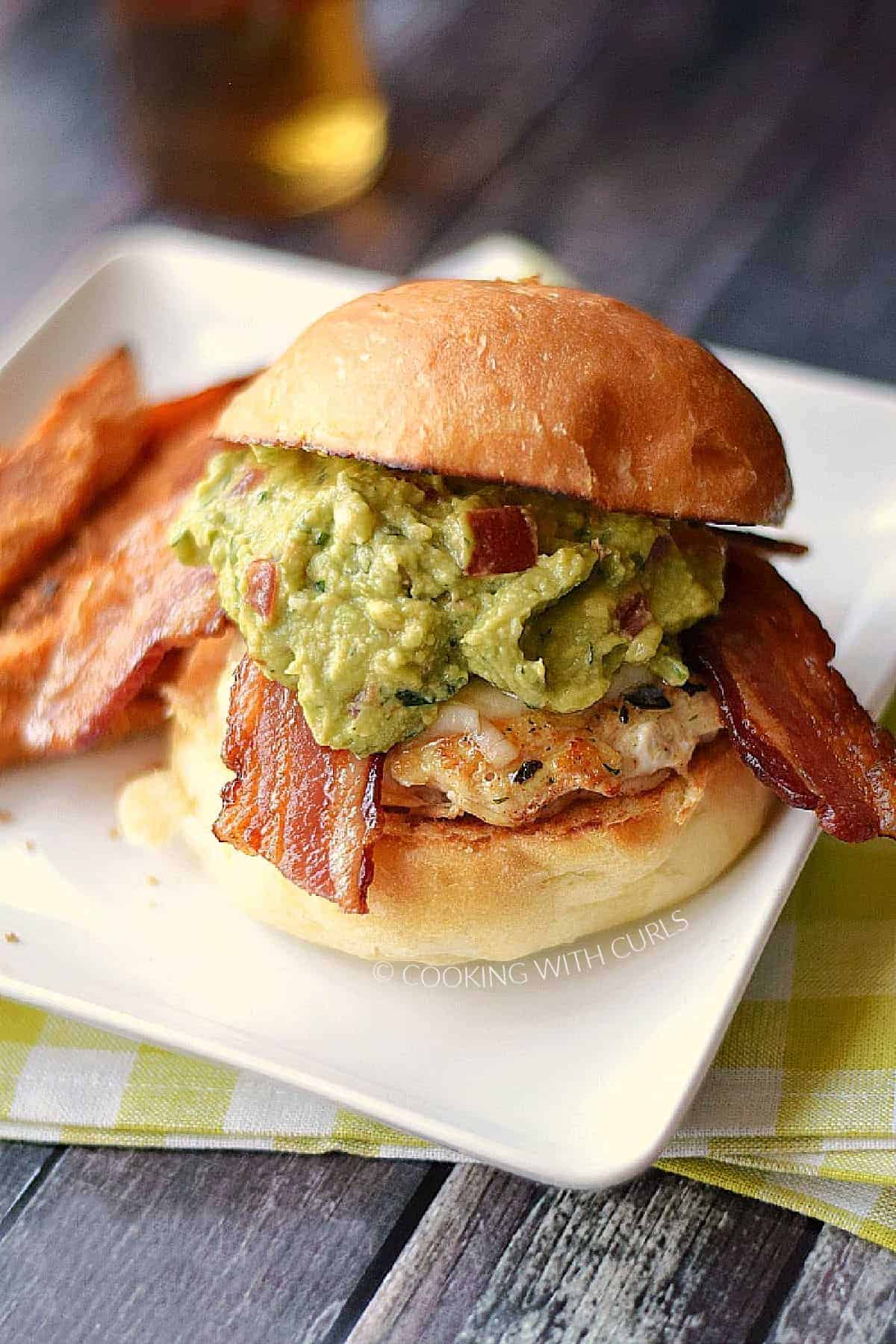 Chicken breast topped with melted cheese, bacon slices, and guacamole on a hamburger bun and sweet potato fries on the side.