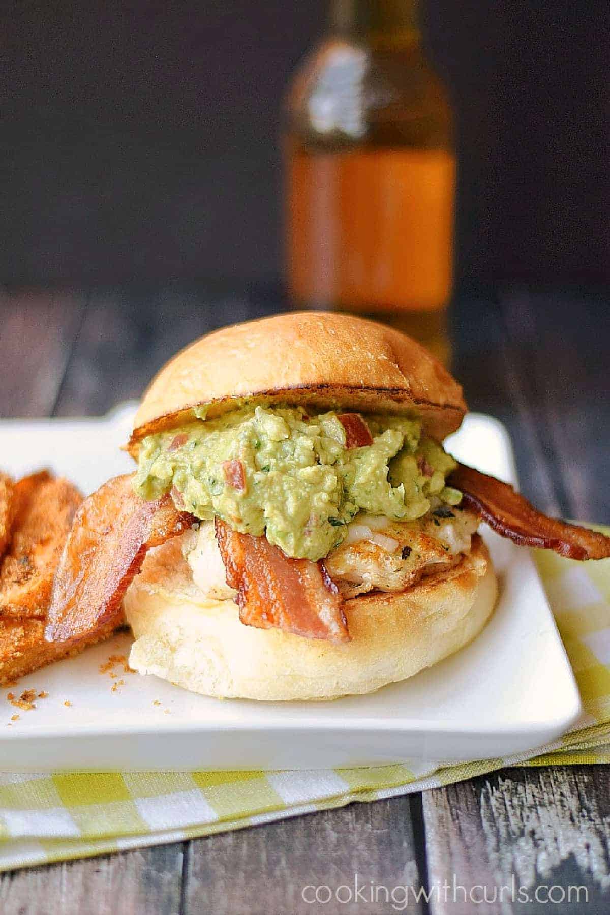 Chicken breast topped with melted cheese, bacon slices, and guacamole on a hamburger bun.