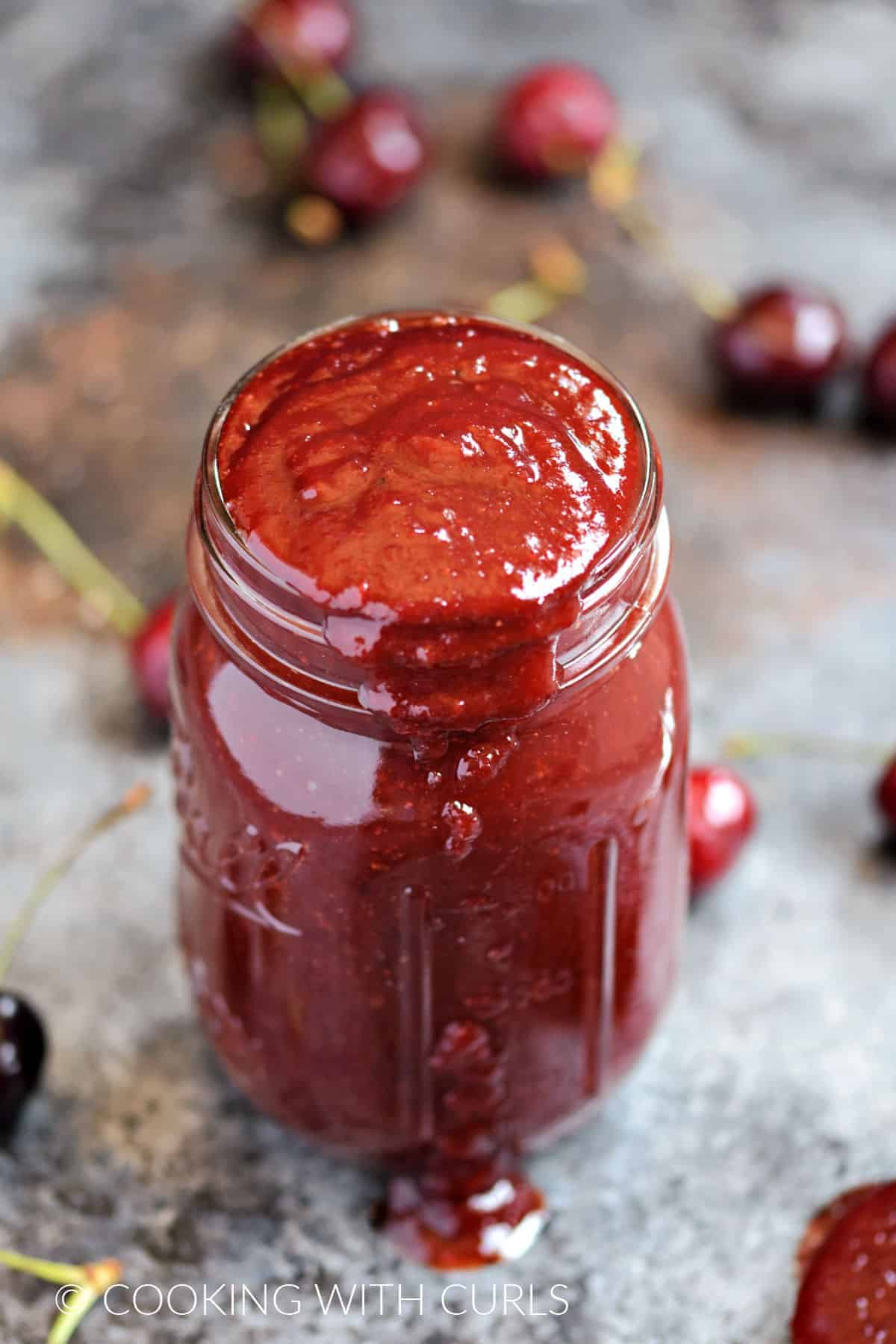 A glass jar overflowing with cherry barbecue sauce surrounded by fresh cherries.
