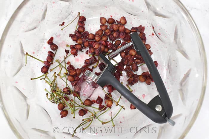 Cherry pits and stems with a cherry pitting tool inside a glass bowl. 