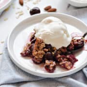 Cherries topped with oat crumble and vanilla ice cream on a small white plate.