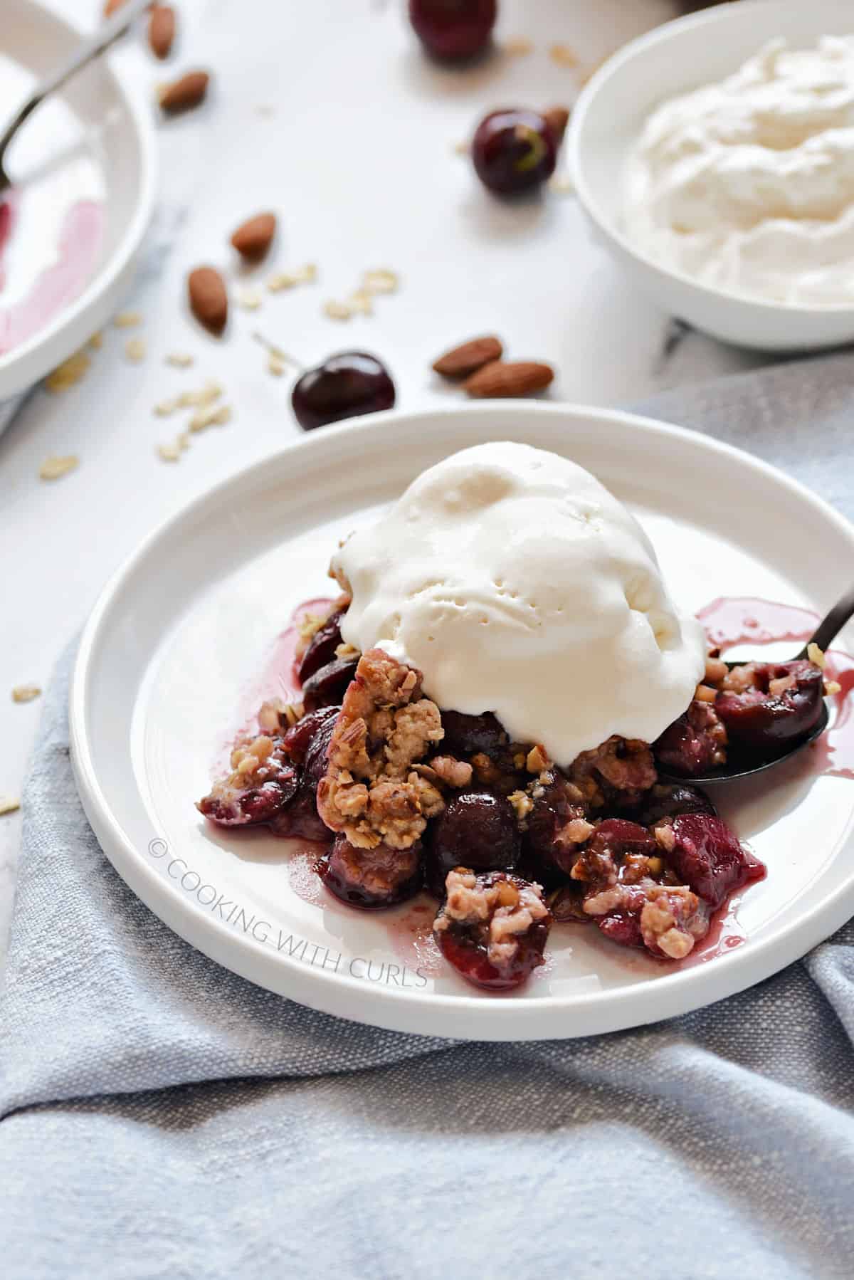 Fresh, baked cherry halves topped with a crunchy oat topping and vanilla ice cream on a small white plate.