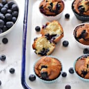 One split and five whole blueberry muffins on a white tray with a bowl of fresh blueberries off to the side.